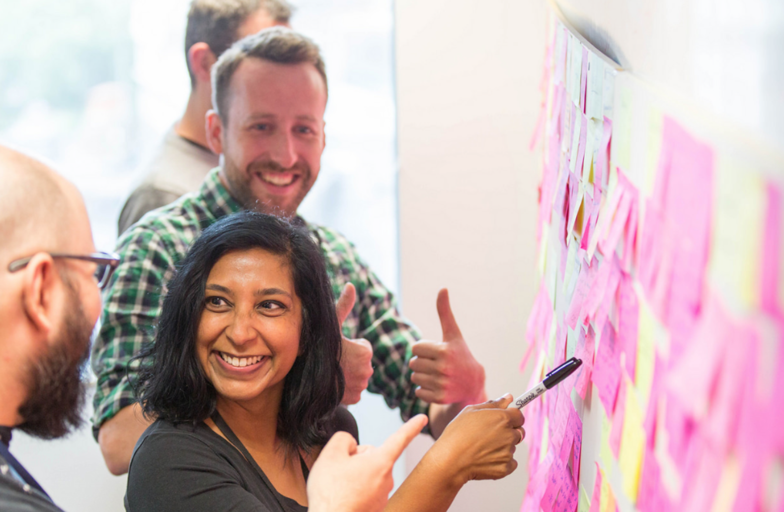 A few people are standing next to a post-it wall smiling and seemingly discussing something