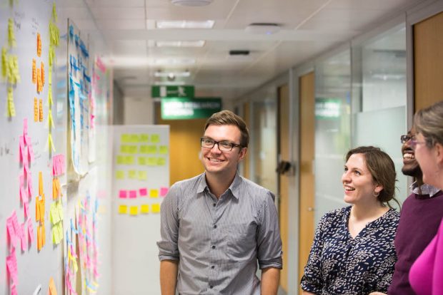Colin Pattinson, Digital, Data and Technology Fast Streamer, standing with his colleagues next to a post-it note wall and smiling