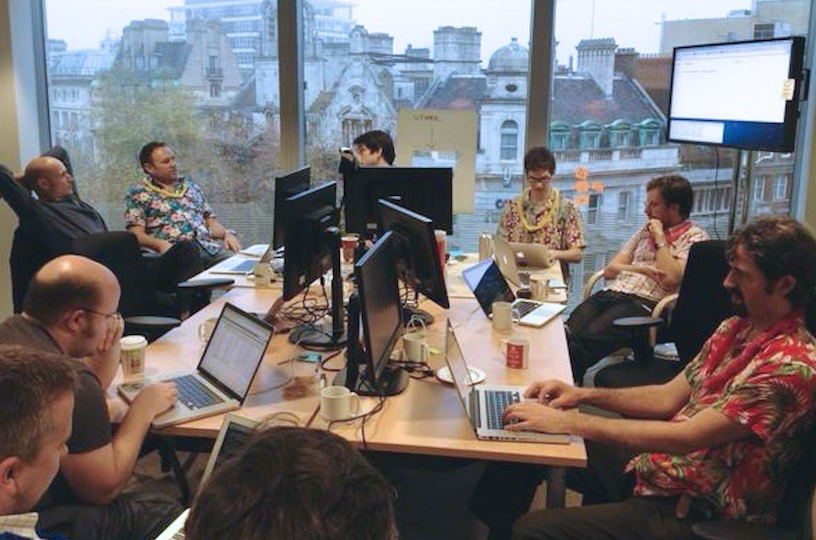 Colleagues in the office environment at GDS in 2012, sitting around the square table, next to computer screens and on laptops.