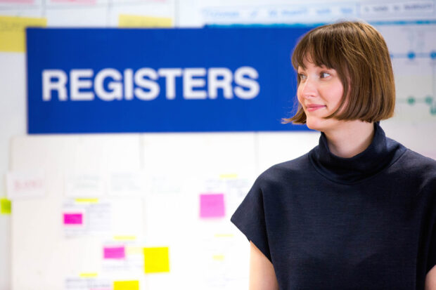 Michaela standing in front of a camera next to a sign that reads "Registers"