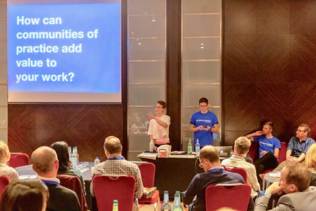 An audience watching a conference presentation. On the screen are the words "How can communities of practice add value to your work?"