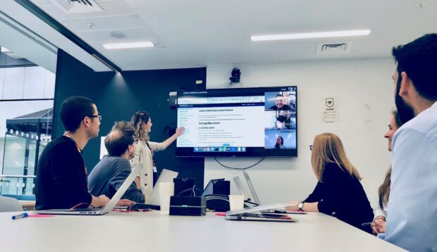 A team of coworkers sit at a table in a meeting room, looking while a woman points out some text on a screen