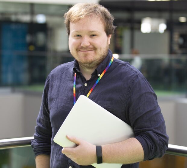 Lewis Dunne, holding a laptop and standing in an office building