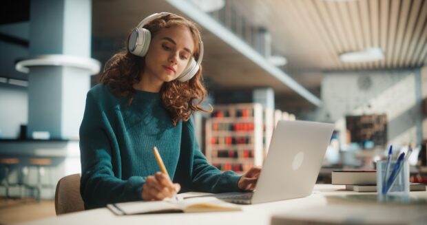 Concentrated Young Female Student Engaged in Academic Research With Laptop and Headphones at Modern Library Desk, Writing Notes and Studying Online.